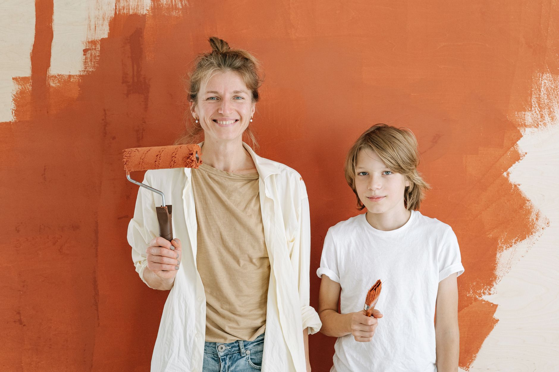 A mother and son smiling while painting a wall indoors, holding paint roller and brush in Temecula, CA